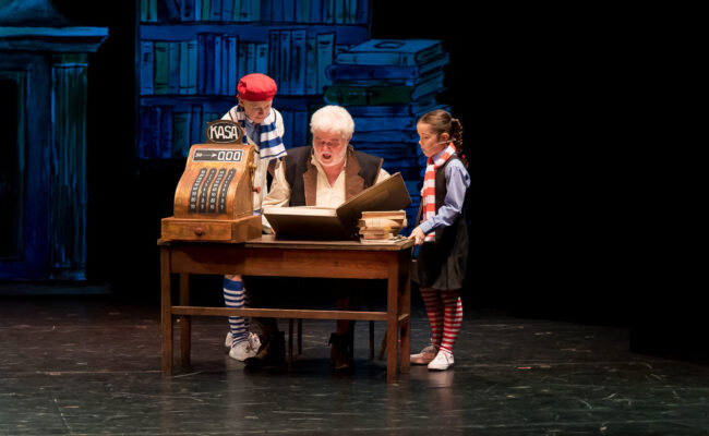 Zdjęcie ilustruje scenę gdzie Kurzugóra siedzi za biurkiem i czyta z wielkiej księgi. Obok niego soją Maks i Roma.